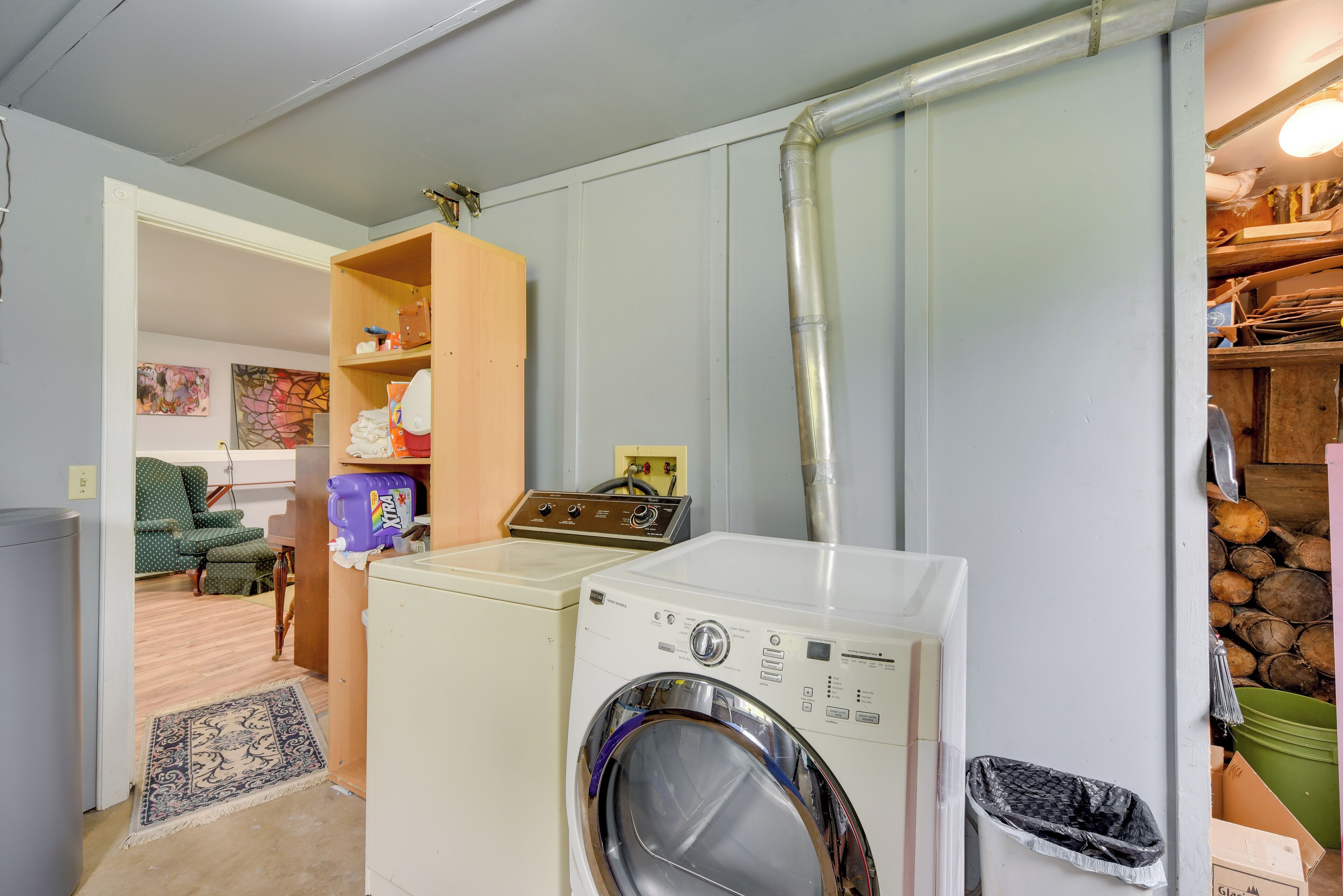 Une buanderie équipée d’un lave-linge et d’un sèche-linge à chargement frontal, d’une étagère en bois sur laquelle sont disposés divers objets, d’un panier à linge violet et d’un seau vert.