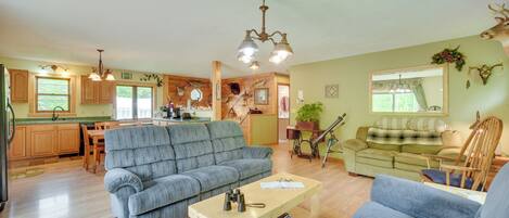 A spacious living room with a grey sofa, a wooden coffee table, and a green sofa. There is a kitchen area with wooden cabinets and a dining table.