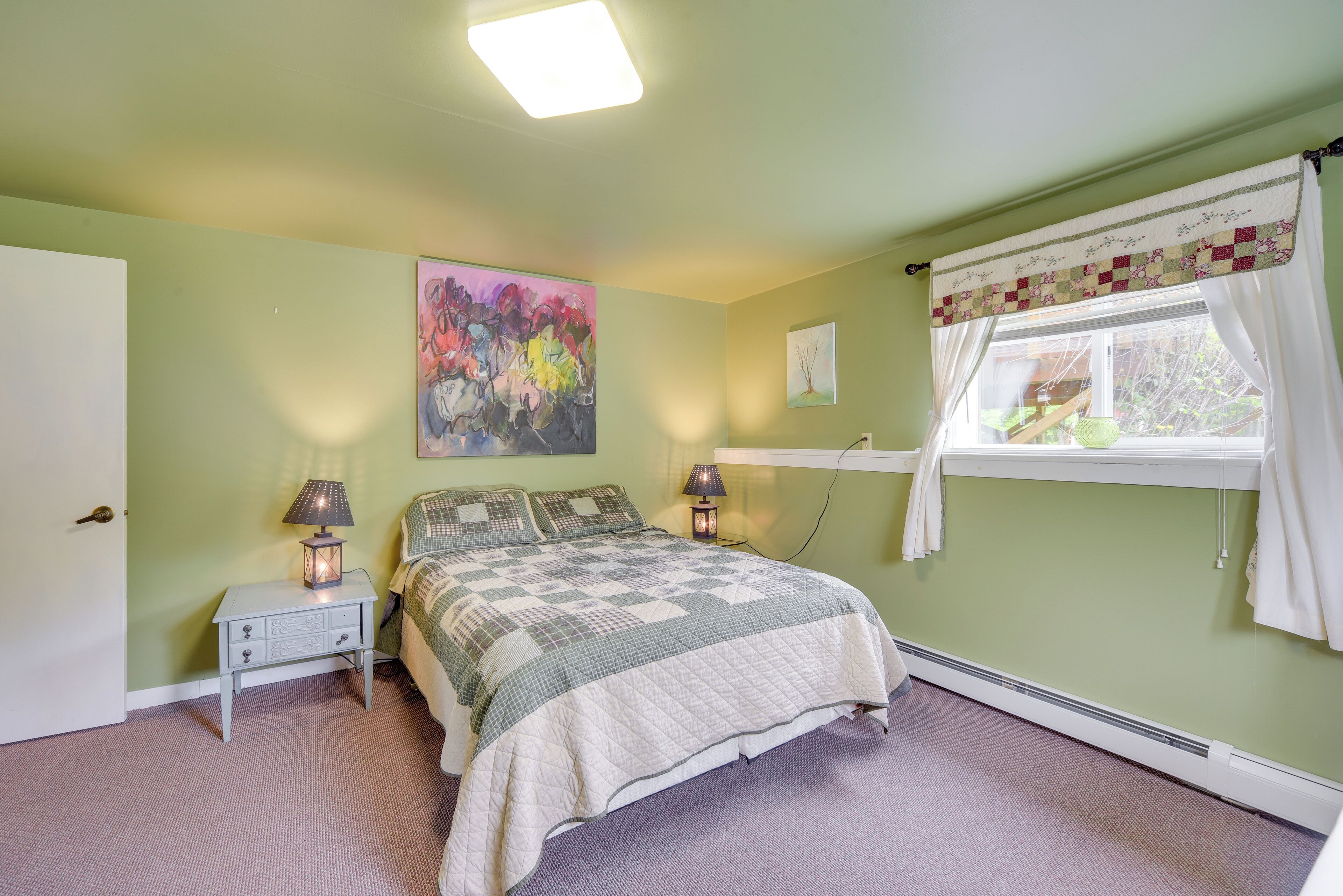 Un dormitorio con cama, mesitas de noche, lámpara, un cuadro y ventana con cortinas.