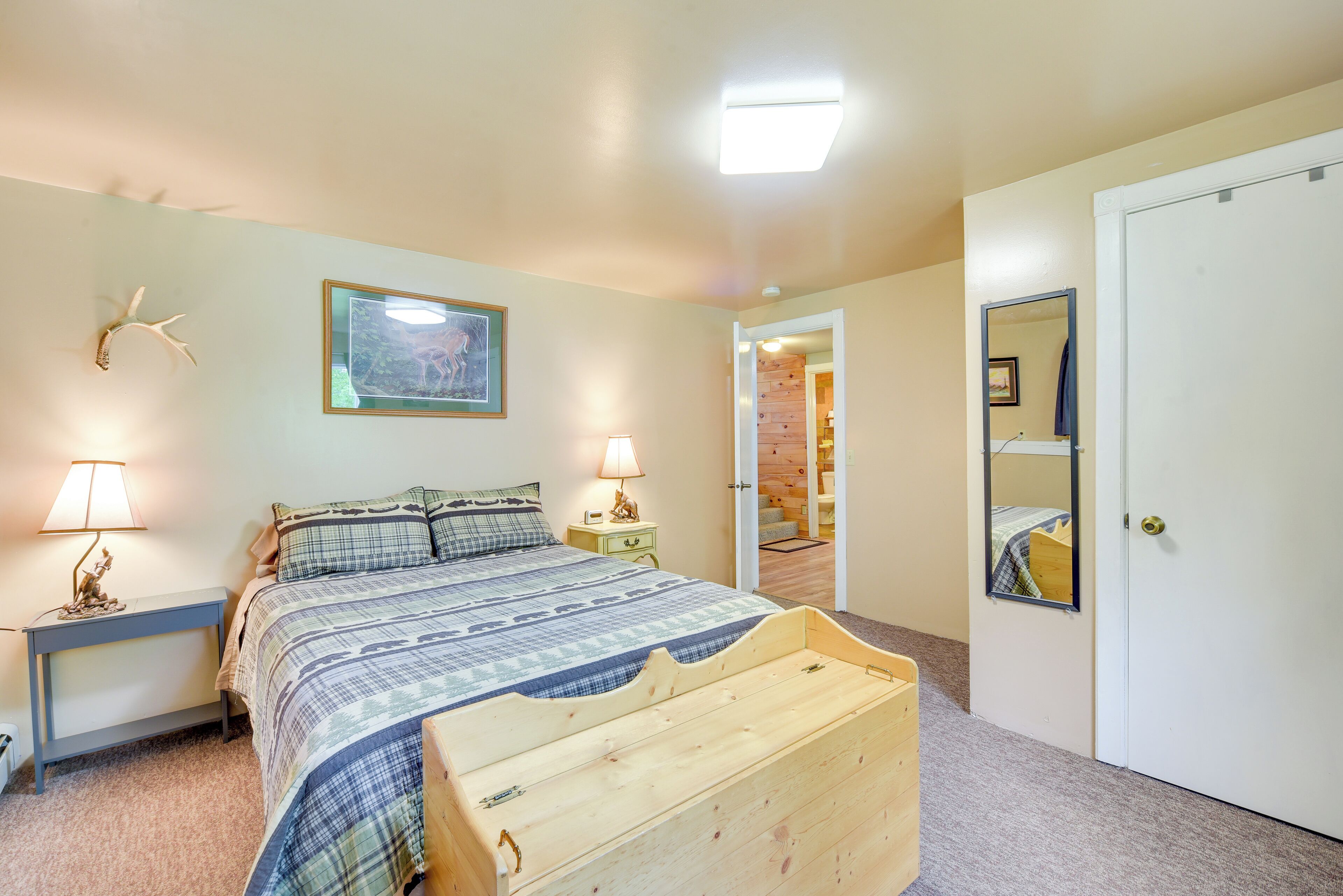 A bedroom with a bed, bedside lamps, a framed picture, a mirror, and a door leading to another room.