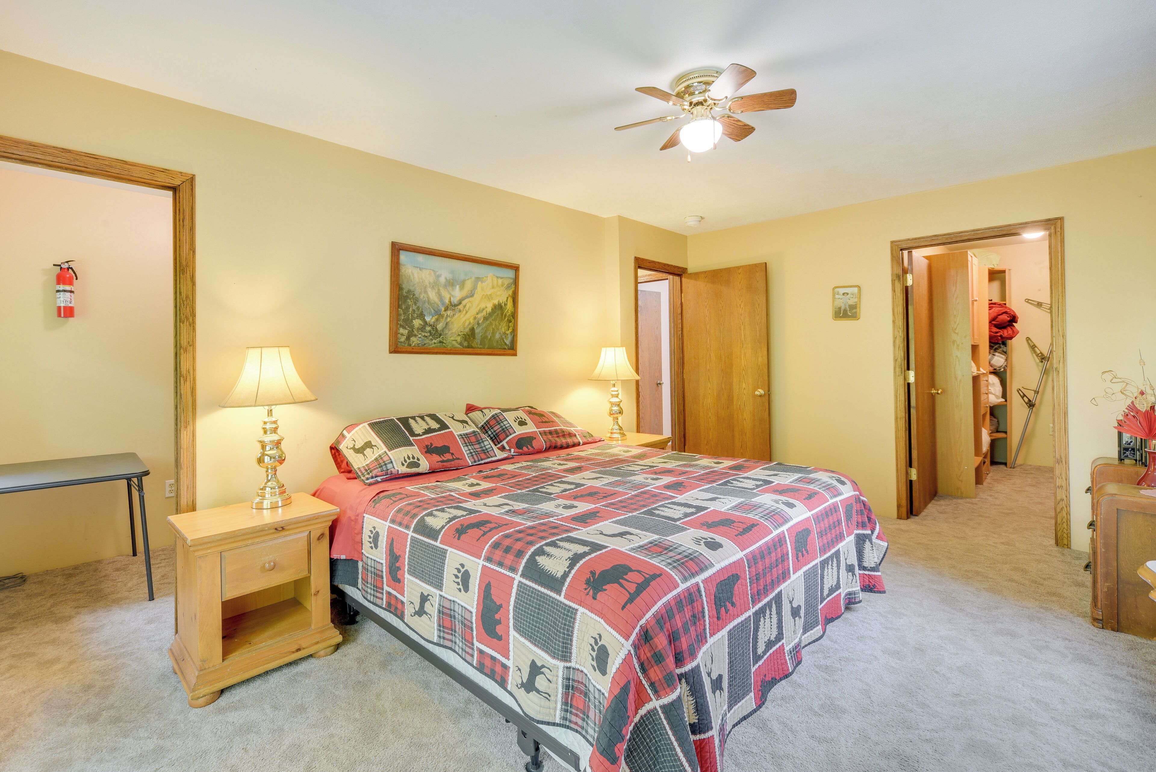A bedroom with a bed, bedside lamps, a ceiling fan, a door, and a fire extinguisher.