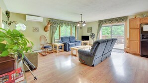 Ein geräumiges Wohnzimmer mit einem blauen Sofa, einem Holztisch und einem blauen Sessel. Ein Fernseher steht auf einem Schrank, daneben ein Zimmerpflanze und ein Fenster mit Vorhängen.