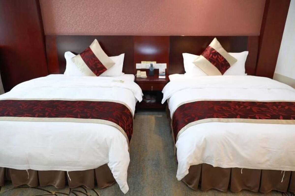 A hotel room with two beds, each with white bedding and red accents, a wooden headboard, and a nightstand with a lamp and phone.
