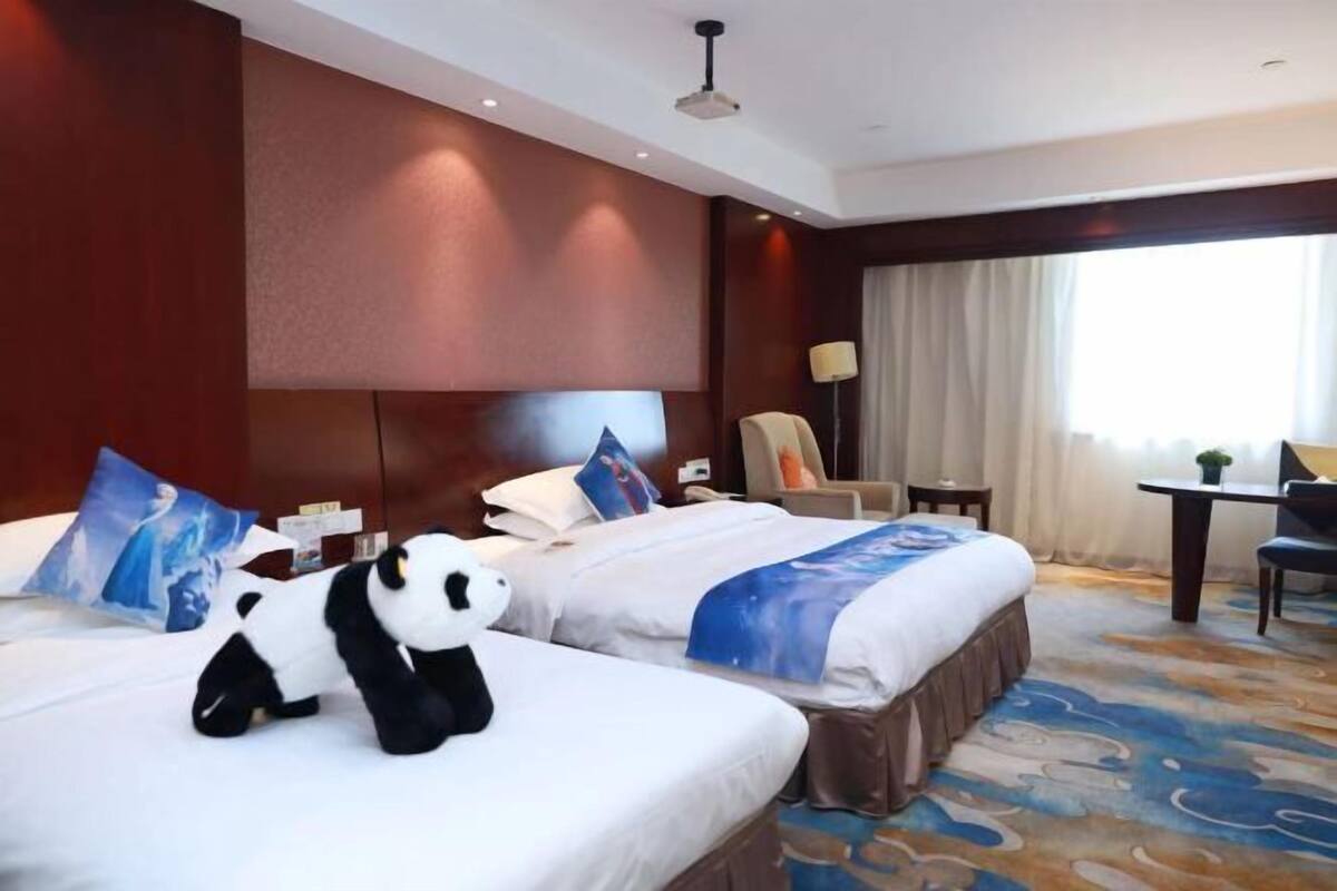 A hotel room with two beds, a desk, a chair, and a panda plush toy.