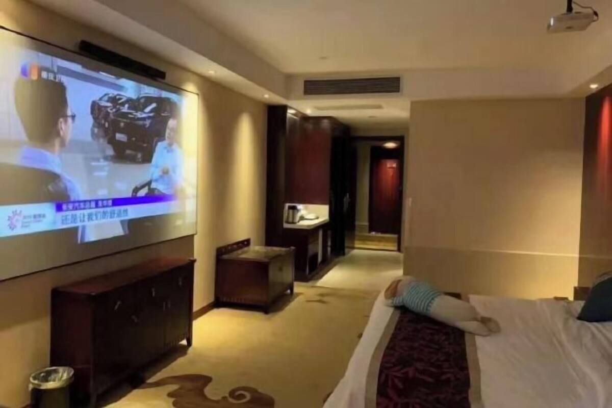 A hotel room with a large flat-screen TV mounted on the wall, a bed with a patterned bedspread, a small wooden side table, and a dark-colored cabinet.