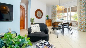 Un salon moderne avec un canapé, une table à manger et des chaises. Un téléviseur est fixé au mur, il y a un miroir rond et une fenêtre avec des rideaux.