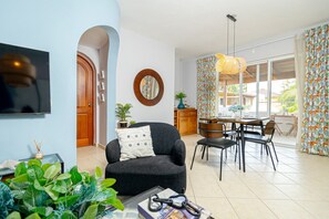 Un salon moderne avec un canapé, une table à manger et des chaises. Un téléviseur est fixé au mur, il y a un miroir rond et une fenêtre avec des rideaux.