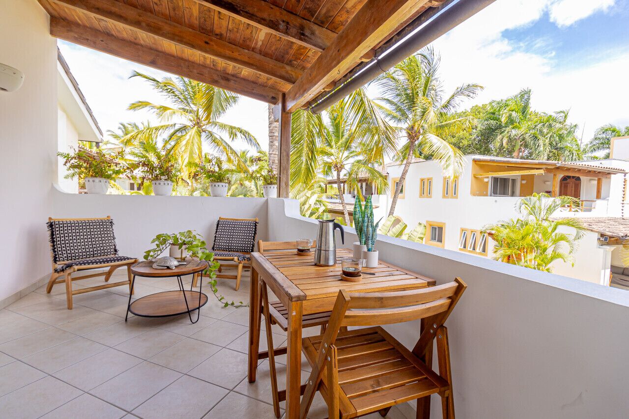 A balcony with wooden furniture, potted plants, and a view of palm trees and buildings.