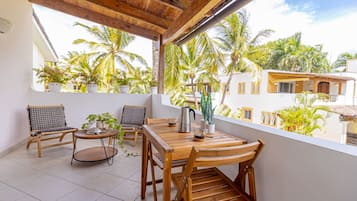 A balcony with wooden furniture, potted plants, and a view of palm trees and buildings.