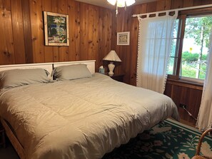 Une chambre à coucher aux murs lambrissés, avec un lit, un tableau encadré, une lampe et une fenêtre avec des rideaux.