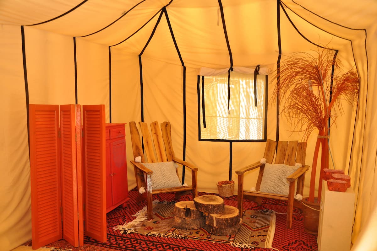 Una carpa con dos sillas de madera, un tapete tejido y una ventana con cortinas.