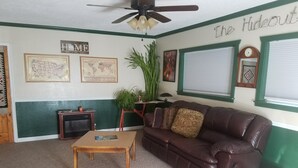 A living room with a brown leather sofa, a wooden coffee table, a ceiling fan, a map on the wall, and a window with blinds. - The Lazy Ass Ranch. The National Parks Room (Panguitch)