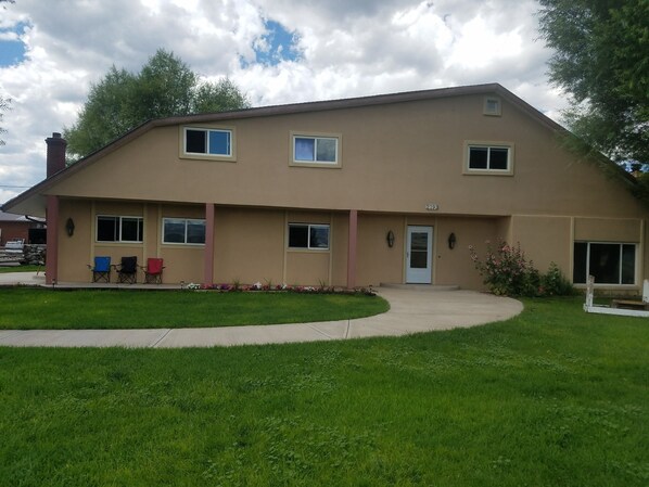 A two-story building with a beige exterior, numbered 2193, featuring a front porch with outdoor seating and a well-maintained lawn. - The Lazy Ass Ranch. The National Parks Room (Panguitch)