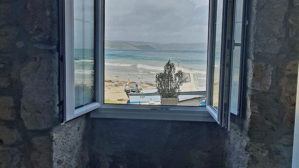 Une pièce avec un mur en pierre et une fenêtre donnant sur une plage où l’on aperçoit un bateau et une plante.