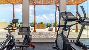 A gym with exercise equipment, a digital screen, and a view of the sky and clouds through large windows.