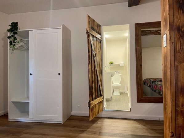 A room with a white wardrobe, a wooden door, a bathroom with a toilet and sink, and a mirror on the wall. - Jaune Hôtel Boutique (Saint-Gédéon)