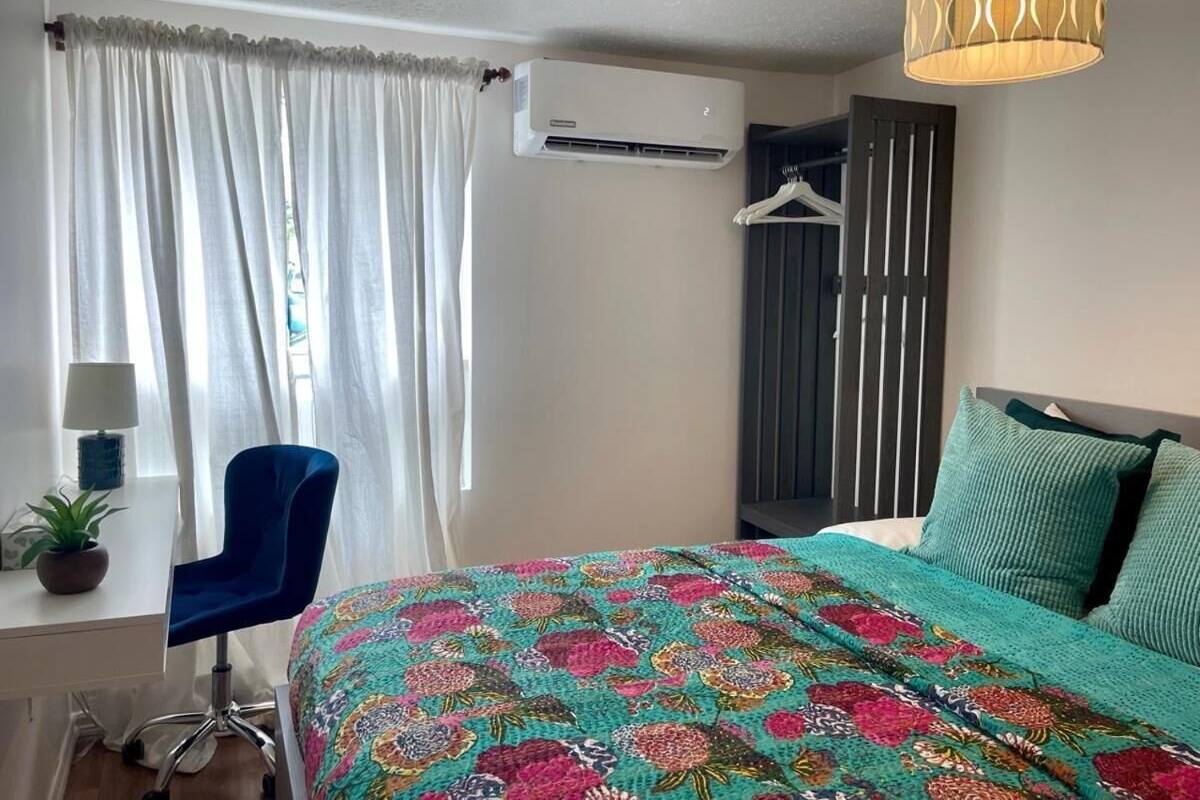 A bedroom with a bed, a desk, a chair, an air conditioning unit, and a hanging light fixture.
