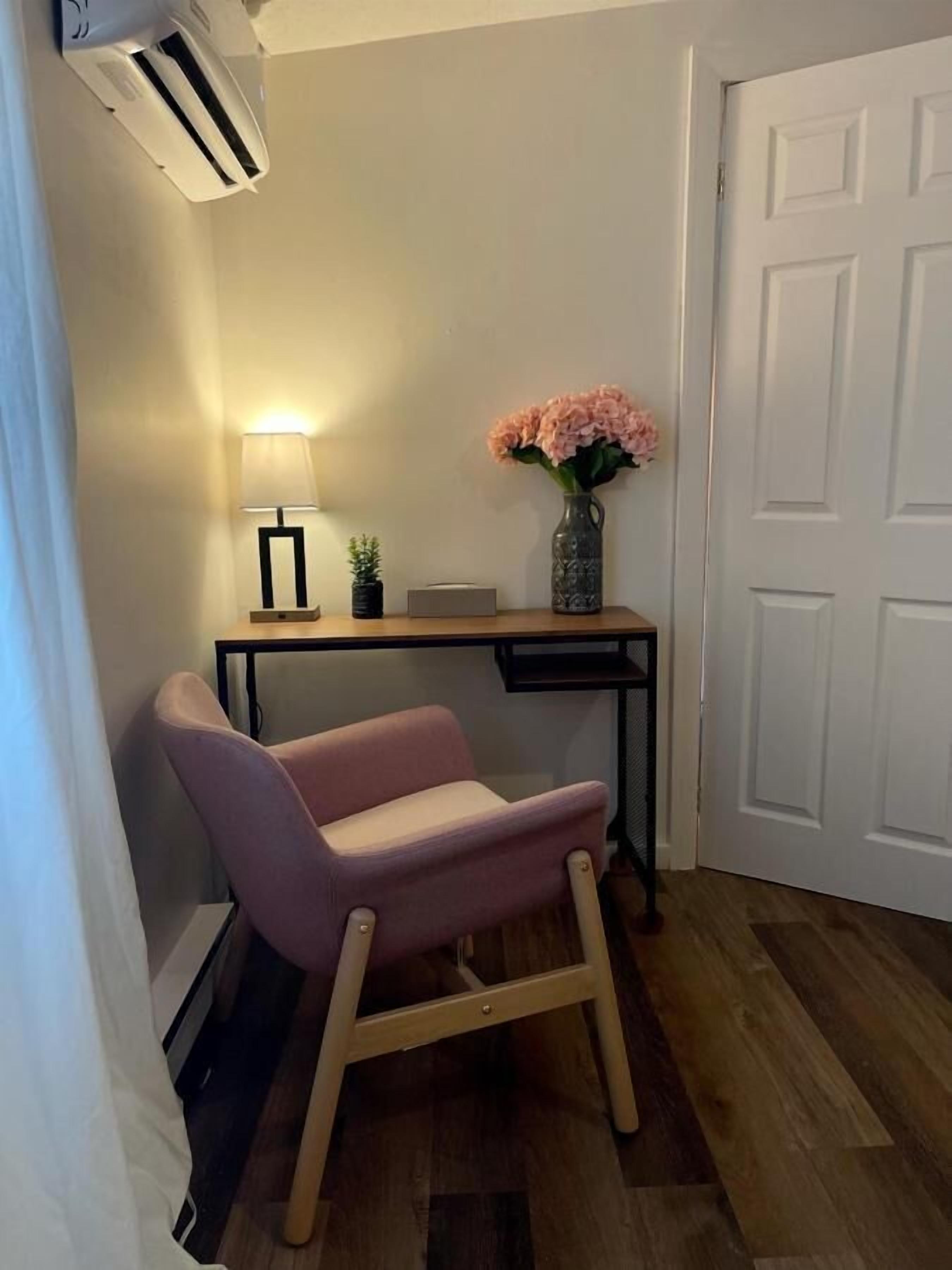 A corner room with a wooden desk, a pink chair, a lamp, a vase with flowers, and a white door.