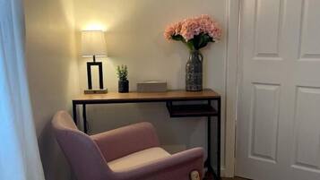 A corner room with a wooden desk, a pink chair, a lamp, a vase with flowers, and a white door.