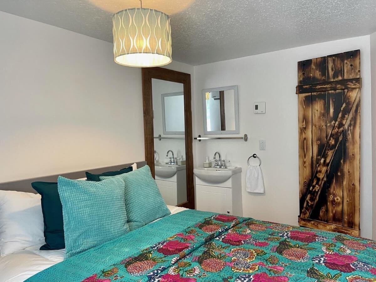 A bedroom with a bed, a bedside table, a mirror, a sink, and a wooden door.