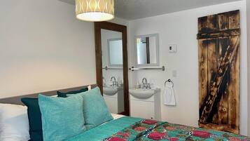 A bedroom with a bed, a bedside table, a mirror, a sink, and a wooden door.