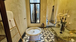 Un baño con suelo de baldosas blanco y negro, un lavabo de vidrio, un banco de madera y una ventana con cortinas.
