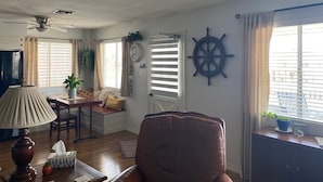A living room with a ceiling fan, a dining area with a table and chairs, a brown leather chair, a ship's wheel decoration, and a window with blinds. - Charming 2-bedroom waterfront house with dock, AC, and WiFi in Rockport Texas (Rockport)