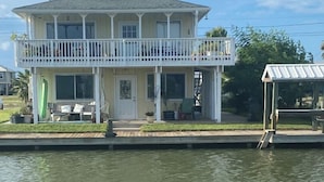 A two-story house with a balcony, a dock extending into a body of water, and a small covered structure to the right. - Charming 2-bedroom waterfront house with dock, AC, and WiFi in Rockport Texas (Rockport)