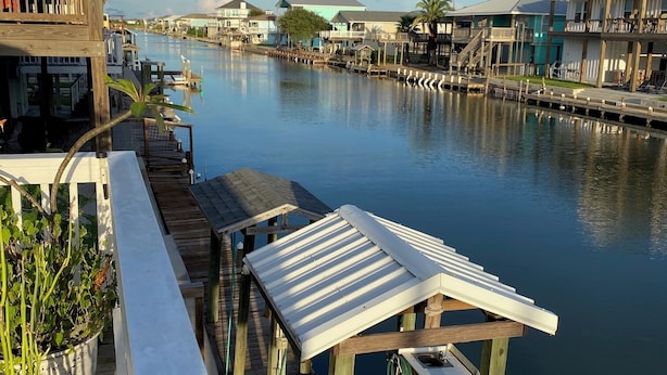 A wooden pier extending over calm water, with a row of tables and benches, potted plants, and a view of waterfront buildings. - Charming 2-bedroom waterfront house with dock, AC, and WiFi in Rockport Texas (Rockport)