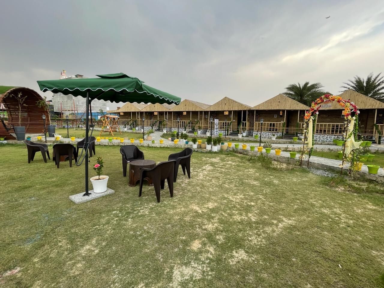 An outdoor seating area with tables and chairs, surrounded by a grassy lawn and a wooden structure in the background.