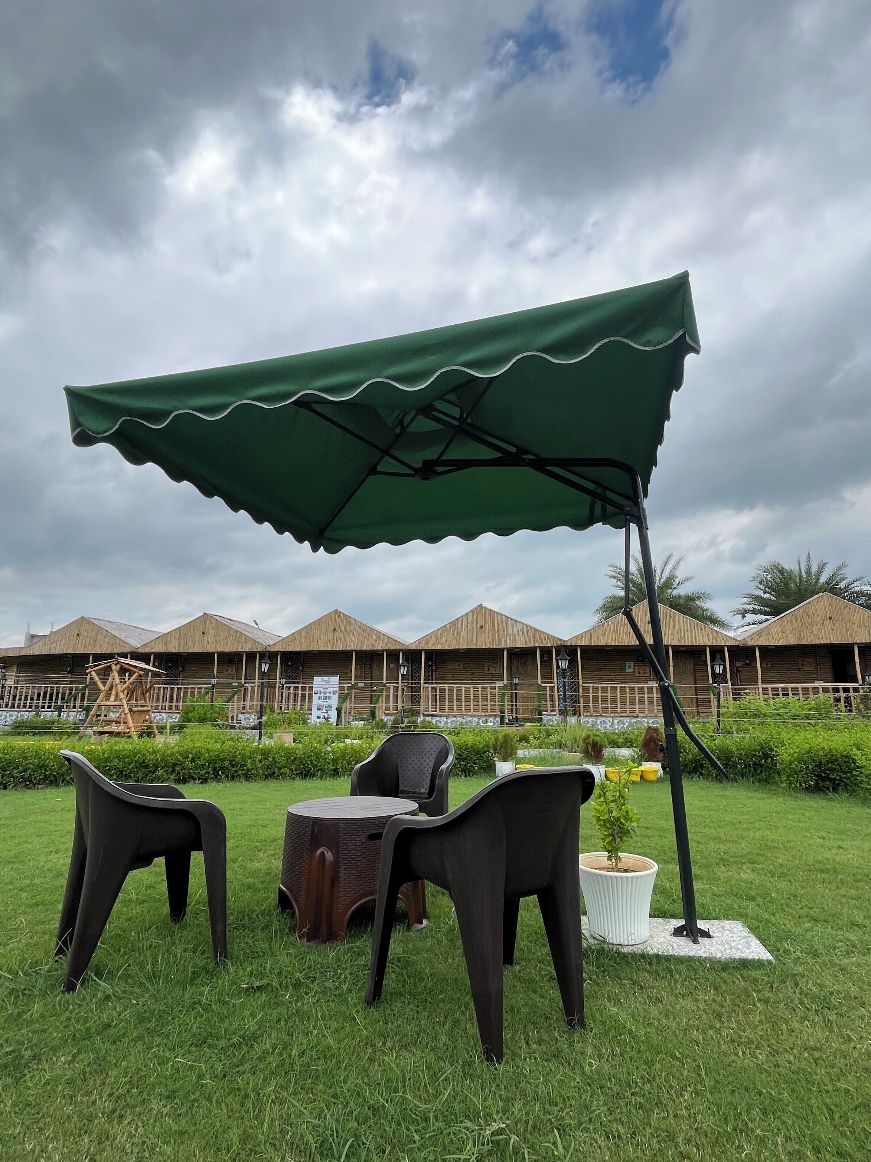 A grassy outdoor area with a green canopy, two wicker chairs, a small round table, and a potted plant. In the background, there are wooden buildings with a thatched roof.