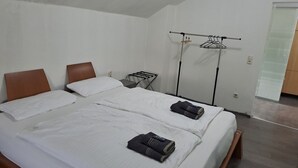 Una cama doble con ropa de cama blanca, un cabecero de madera y una mesita de noche pequeña. Se observa un tendedero de pared con ropa y un espejo.