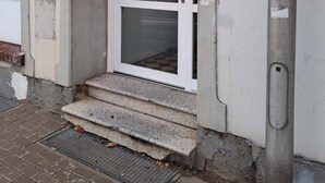 Entrada de un edificio con una puerta blanca, unos pocos escalones y un poste de metal con un letrero.