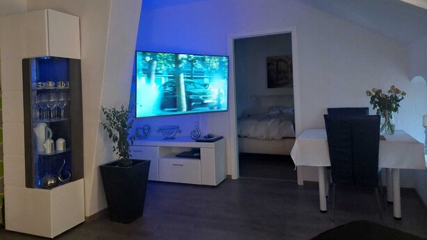 A modern living room with a flat-screen TV, a glass cabinet with glasses, a potted plant, and a dining area with a table and chairs. - Modernes Apartment in Minden am Hauptbahnhof (Minden)
