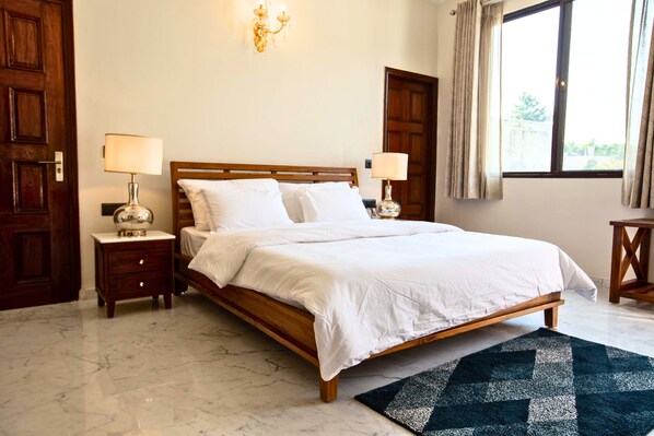 Un dormitorio con cama de madera, dos mesitas de noche con lámparas, suelo de mármol y una ventana con cortinas.