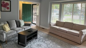 Una sala de estar con un sofá gris, un sofá color beige, una mesa de centro negra y un tapete estampado. Hay una puerta verde y una ventana con persianas.