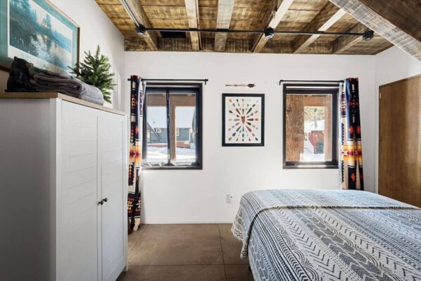 Un dormitorio con cama, dos ventanas, un armario y un cuadro en la pared.