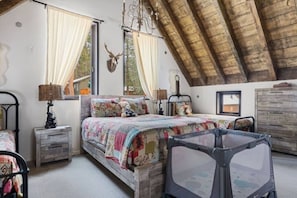 A bedroom with a sloped ceiling, a bed with a colorful quilt, a deer head decoration, and a window with curtains.