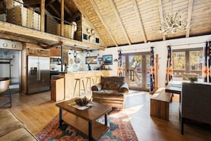 A spacious living area with a high ceiling, wooden beams, a kitchen with a stainless steel refrigerator, a dining area with a bench, and a large window with curtains.
