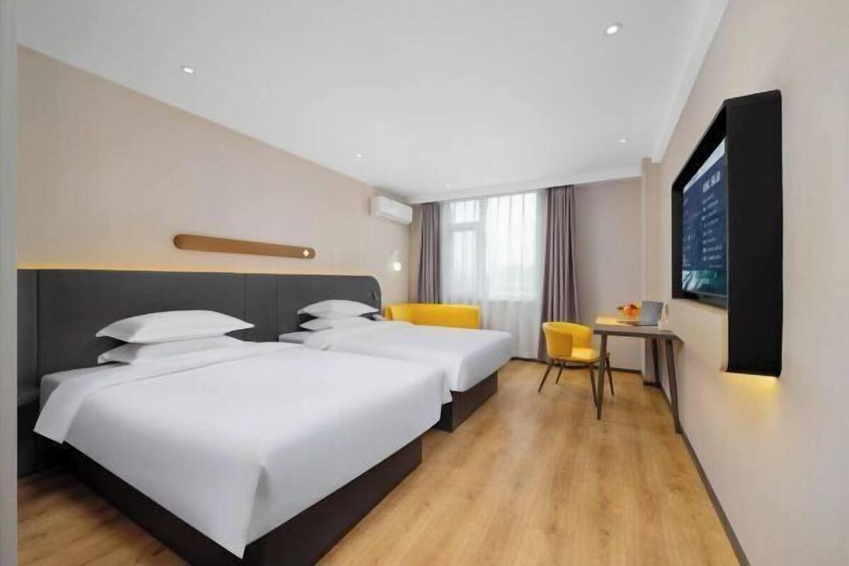 A hotel room with two beds, a small table, a chair, a TV, and a window with curtains.