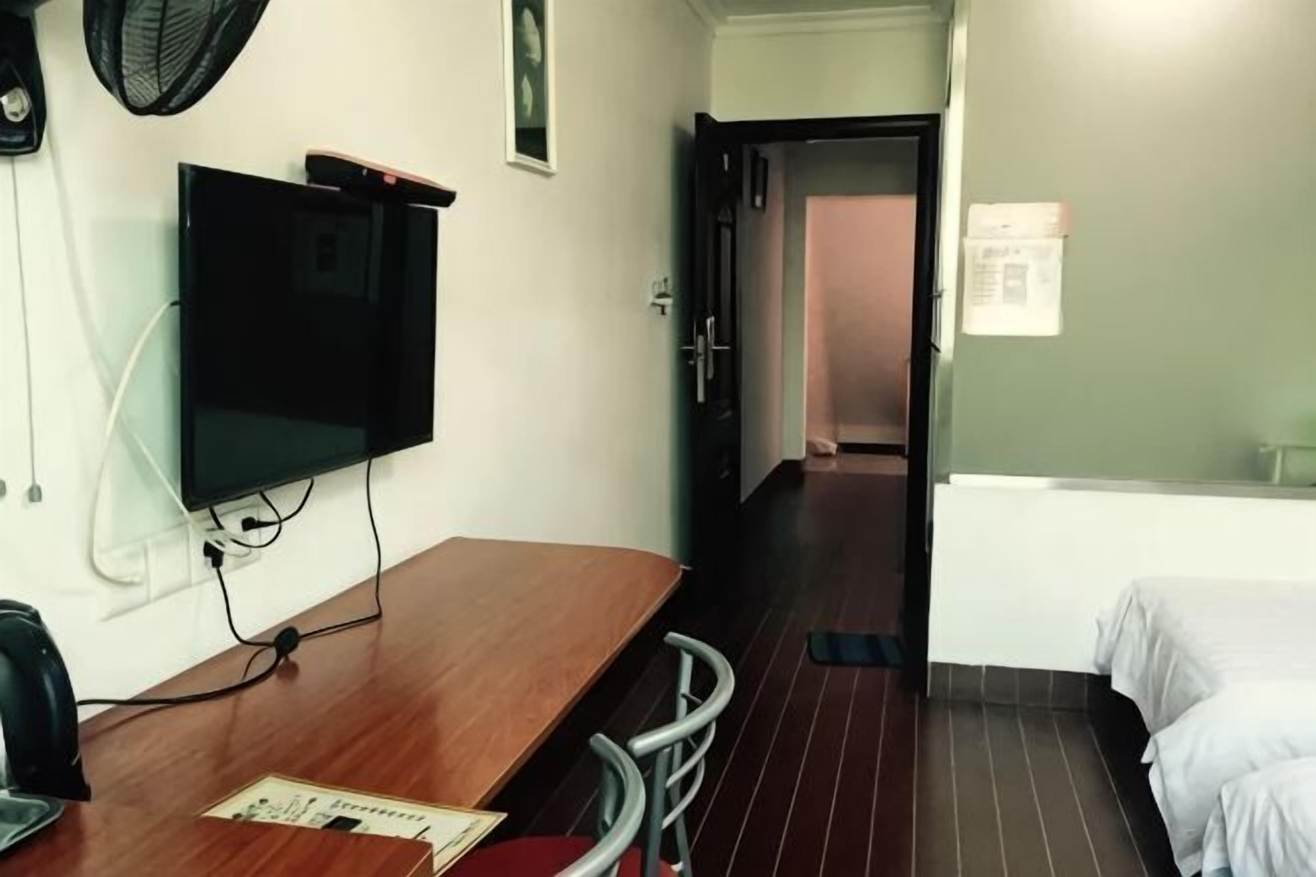 A hotel room with a wooden desk, a television, a bed with white bedding, and a door leading to another room.