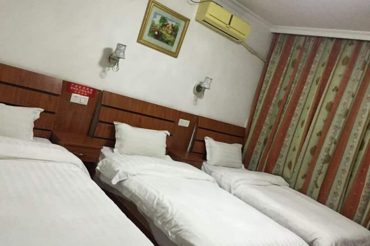 A hotel room with three single beds, a wooden headboard, an air conditioning unit, a wall-mounted clock, and a curtain with a floral pattern.
