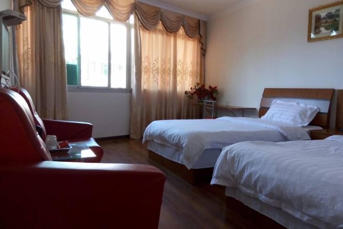 A hotel room with two beds, a red armchair, a small table with a vase of flowers, and a window with curtains.