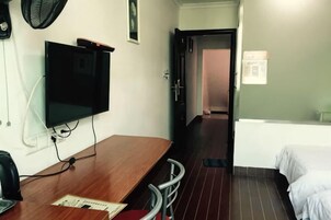 Quarto de hotel com escrivaninha de madeira, televisão, cama e porta para outro espaço.