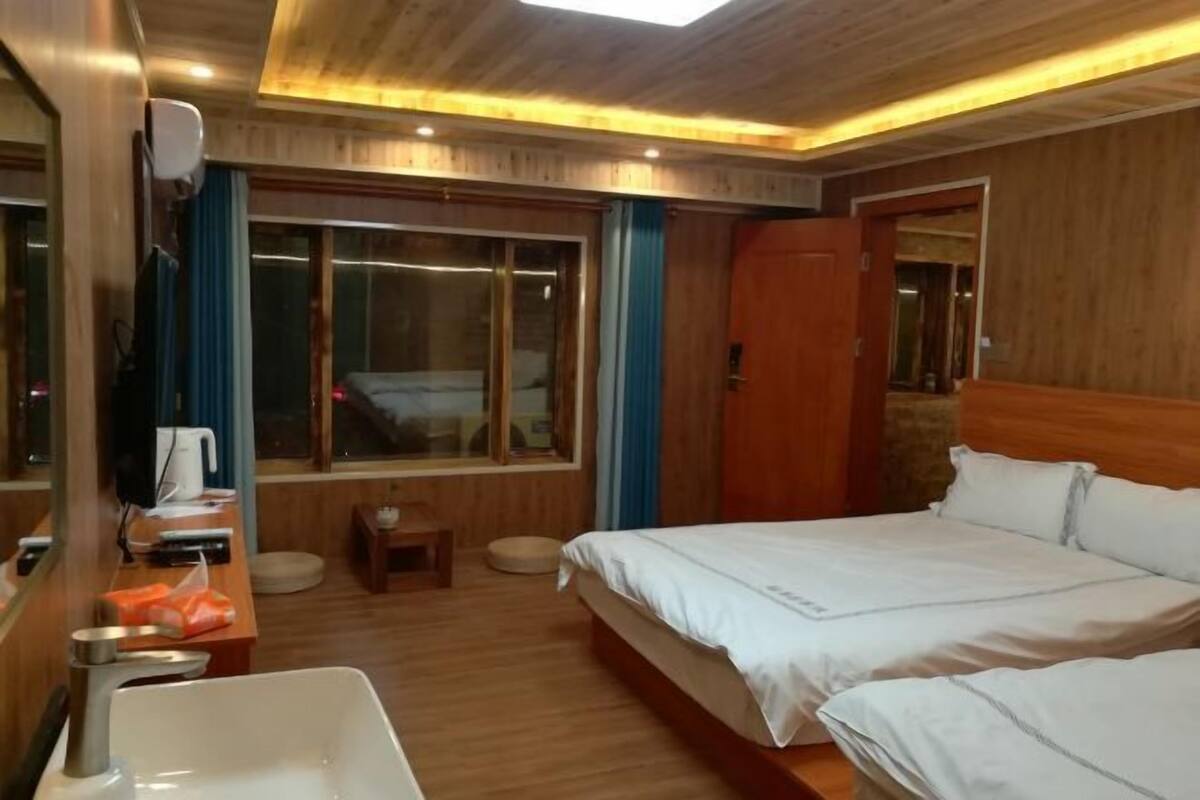 A hotel room with a wooden ceiling, a large window, a bed, a bedside table, and a mirror.