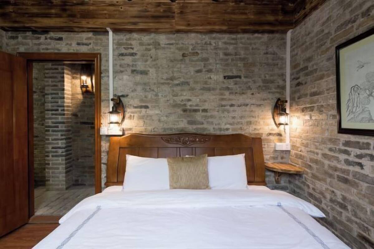 A room with a brick wall, a wooden ceiling, a bed with white bedding, and two wall-mounted lights.