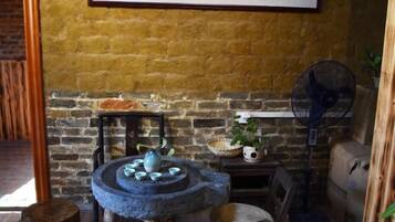Une pièce rustique avec un mur en briques, une table ronde en pierre surmontée de poteries bleues, une petite étagère avec une plante et un ventilateur sur pied.