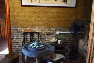 Una habitación rústica con pared de ladrillo, una mesa redonda de piedra con cerámica azul, un estante pequeño con una planta y un ventilador de pie.