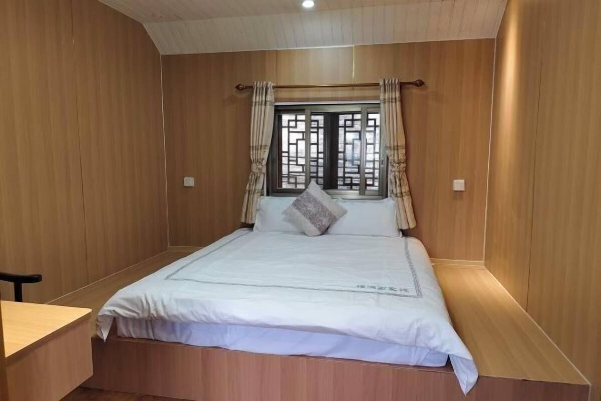 A single bed with a white bedspread and two pillows, a wooden desk, and a window with curtains.