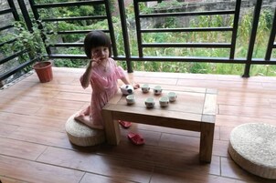 Un niño sentado a una mesa de madera con tazas de té, en una terraza de madera con plantas en macetas y vista a follaje verde.